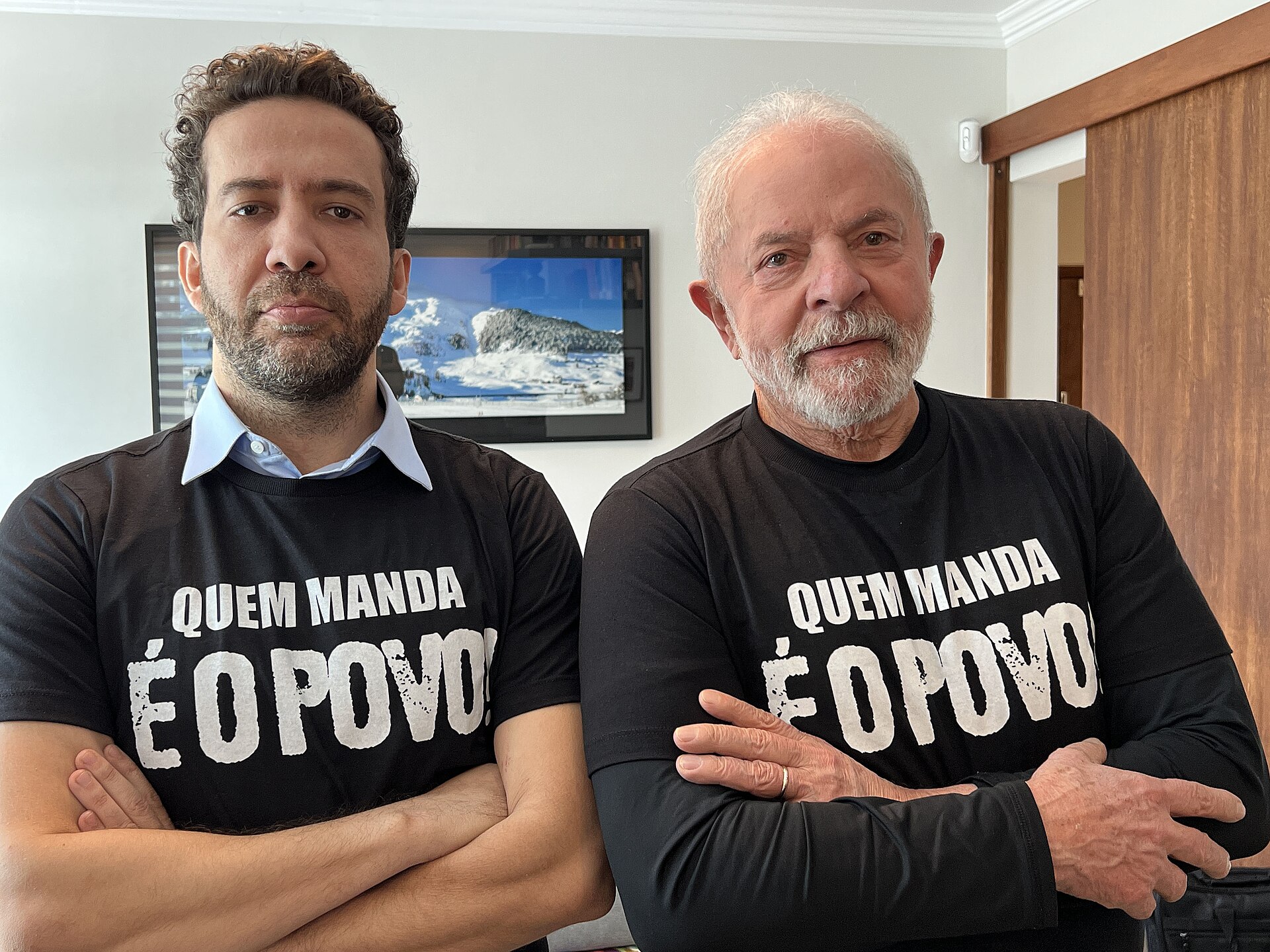 André Janones e Lula lado a lado, de braços cruzados, usando camisetas pretas com a frase "Quem manda é o povo"